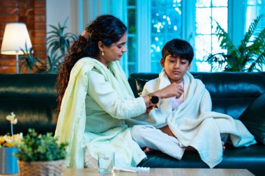 Hintli anne oğlunun ateşini kontrol ederken kanepenin üzerinde oturuyor, sağlığıyla ilgileniyor, sevgisini, sevgisini, sevgisini, aile bağlarını, evde aile yaşam tarzını, günlük rutini besliyor.