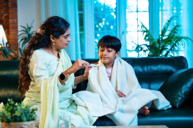 Hintli anne oğlunun ateşini kontrol ederken kanepenin üzerinde oturuyor, sağlığıyla ilgileniyor, sevgisini, sevgisini, sevgisini, aile bağlarını, evde aile yaşam tarzını, günlük rutini besliyor.