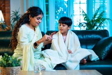 Hintli anne oğlunun ateşini kontrol ederken kanepenin üzerinde oturuyor, sağlığıyla ilgileniyor, sevgisini, sevgisini, sevgisini, aile bağlarını, evde aile yaşam tarzını, günlük rutini besliyor.