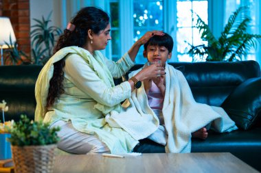 Hintli anne oğlunun ateşini kontrol ederken kanepenin üzerinde oturuyor, sağlığıyla ilgileniyor, sevgisini, sevgisini, sevgisini, aile bağlarını, evde aile yaşam tarzını, günlük rutini besliyor.