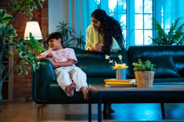 Hintli anne oğlunu azarlıyor, modern evde koltukta otururken, iyi alışkanlıklar öğretiyor, disiplini teşvik ediyor, kaliteli zaman geçirtiyor, çocuğa sevgiyle yol gösteriyor, ebeveyn çocuk ilişkisini kapalı alanda gösteriyor.