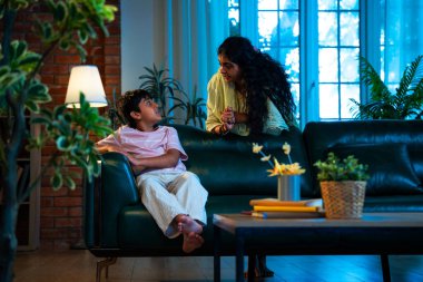 Hintli anne oğlunu azarlıyor, modern evde koltukta otururken, iyi alışkanlıklar öğretiyor, disiplini teşvik ediyor, kaliteli zaman geçirtiyor, çocuğa sevgiyle yol gösteriyor, ebeveyn çocuk ilişkisini kapalı alanda gösteriyor.