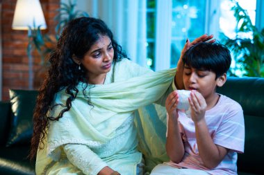 Hintli küçük bir çocuk modern bir evde annesinin yanında otururken öksürüyor, viral semptomlar gösteriyor, kendini iyi hissetmiyor, annesinin bakımı, çocuk bakımı, aile bağı, sağlık farkındalığı, bakımı,