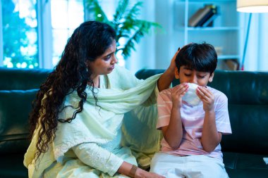 Hintli küçük bir çocuk modern bir evde annesinin yanında otururken öksürüyor, viral semptomlar gösteriyor, kendini iyi hissetmiyor, annesinin bakımı, çocuk bakımı, aile bağı, sağlık farkındalığı, bakımı,