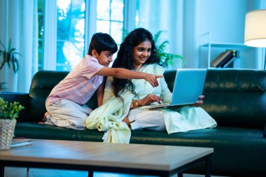 Hintli küçük çocuk ve anne internette sörf yapıyor ya da internetten alışveriş yapıyor modern evdeki kanepede otururken, birlikte kaliteli zaman geçiriyorlar, bağlanma, bağ kurma, ebeveyn çocuk yaşam tarzı gösteriyorlar.