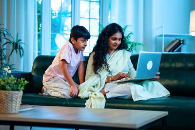 Hintli küçük çocuk ve anne internette sörf yapıyor ya da internetten alışveriş yapıyor modern evdeki kanepede otururken, birlikte kaliteli zaman geçiriyorlar, bağlanma, bağ kurma, ebeveyn çocuk yaşam tarzı gösteriyorlar.