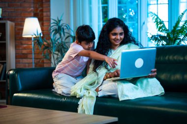 Hintli küçük çocuk ve anne internette sörf yapıyor ya da internetten alışveriş yapıyor modern evdeki kanepede otururken, birlikte kaliteli zaman geçiriyorlar, bağlanma, bağ kurma, ebeveyn çocuk yaşam tarzı gösteriyorlar.