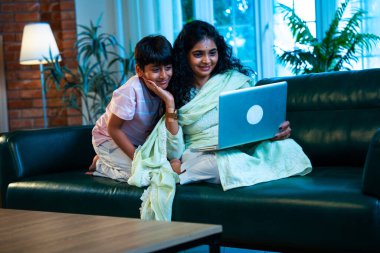 Hintli küçük çocuk ve anne internette sörf yapıyor ya da internetten alışveriş yapıyor modern evdeki kanepede otururken, birlikte kaliteli zaman geçiriyorlar, bağlanma, bağ kurma, ebeveyn çocuk yaşam tarzı gösteriyorlar.