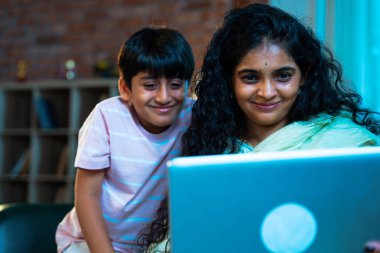 Hintli küçük çocuk ve anne internette sörf yapıyor ya da internetten alışveriş yapıyor modern evdeki kanepede otururken, birlikte kaliteli zaman geçiriyorlar, bağlanma, bağ kurma, ebeveyn çocuk yaşam tarzı gösteriyorlar.