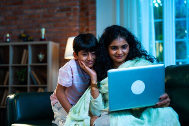 Hintli küçük çocuk ve anne internette sörf yapıyor ya da internetten alışveriş yapıyor modern evdeki kanepede otururken, birlikte kaliteli zaman geçiriyorlar, bağlanma, bağ kurma, ebeveyn çocuk yaşam tarzı gösteriyorlar.