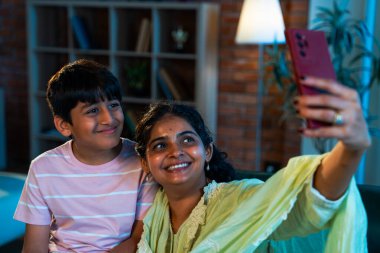 Hintli çocuk, annesiyle selfie çekiyor. Modern evde, kanepenin üzerinde akıllı telefon kullanıyor. Birlikte zaman geçiriyorlar. Şefkat, sevgi, neşe, sıcaklık, aile içi çocuk ilişkileri.