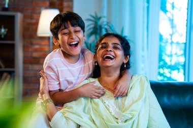 Hintli anne küçük çocuk kanepenin üzerinde modern evde sarılıyor, birlikte iyi vakit geçiriyorlar, sevgi ve sıcaklık gösteriyorlar, ebeveyn-çocuk ilişkisi yaşam tarzı aile mutluluğu kavramı.