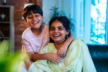 Hintli anne küçük çocuk kanepenin üzerinde modern evde sarılıyor, birlikte iyi vakit geçiriyorlar, sevgi ve sıcaklık gösteriyorlar, ebeveyn-çocuk ilişkisi yaşam tarzı aile mutluluğu kavramı.