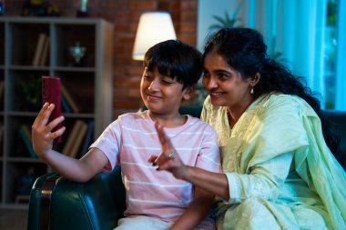 Hintli çocuk, annesiyle selfie çekiyor. Modern evde, kanepenin üzerinde akıllı telefon kullanıyor. Birlikte zaman geçiriyorlar. Şefkat, sevgi, neşe, sıcaklık, aile içi çocuk ilişkileri.