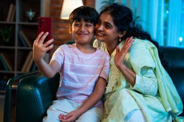 Hintli çocuk ve anne akıllı telefondan video çağrısında bulundular. Modern bir evde, koltukta oturuyorlar, birlikte kaliteli zaman geçiriyorlar. Şefkat, sevgi, aile içi çocuk ilişkisi gösteriyorlar.