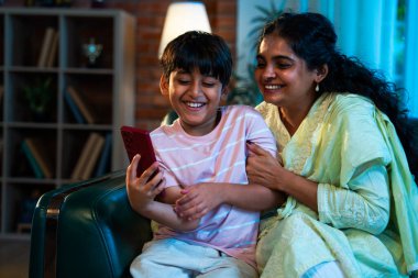 Hintli çocuk ve anne akıllı telefondan sörf yaparken modern evde kanepede oturuyorlar, birlikte kaliteli zaman geçiriyorlar, teknolojiyi kullanıyorlar, neşeyi paylaşıyorlar, ebeveynlik ilişkileri, aile ilişkileri.