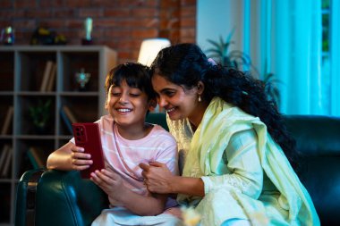Hintli çocuk ve anne akıllı telefondan sörf yaparken modern evde kanepede oturuyorlar, birlikte kaliteli zaman geçiriyorlar, teknolojiyi kullanıyorlar, neşeyi paylaşıyorlar, ebeveynlik ilişkileri, aile ilişkileri.