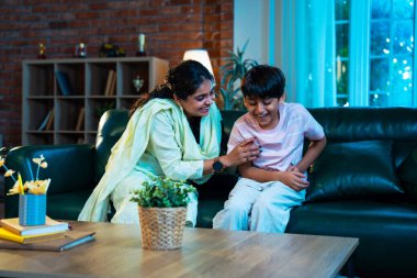 Hintli çocuk ve anne gülerek modern evde kanepede oturuyorlar, birlikte kaliteli zaman geçiriyorlar, şefkat gösteriyorlar, sevgi, neşe, mutluluk, ebeveyn-çocuk ilişkisi yaşam tarzı kapalı