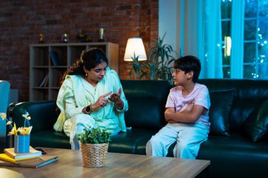 Hintli anne oğlunu azarlıyor, modern evde koltukta otururken, iyi alışkanlıklar öğretiyor, disiplini teşvik ediyor, kaliteli zaman geçirtiyor, çocuğa sevgiyle yol gösteriyor, ebeveyn çocuk ilişkisini kapalı alanda gösteriyor.