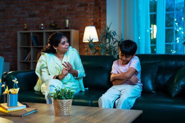 Hintli anne oğlunu azarlıyor, modern evde koltukta otururken, iyi alışkanlıklar öğretiyor, disiplini teşvik ediyor, kaliteli zaman geçirtiyor, çocuğa sevgiyle yol gösteriyor, ebeveyn çocuk ilişkisini kapalı alanda gösteriyor.