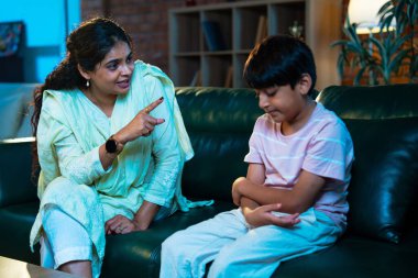 Hintli anne oğlunu azarlıyor, modern evde koltukta otururken, iyi alışkanlıklar öğretiyor, disiplini teşvik ediyor, kaliteli zaman geçirtiyor, çocuğa sevgiyle yol gösteriyor, ebeveyn çocuk ilişkisini kapalı alanda gösteriyor.