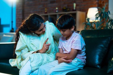 Hintli anne oğlunu azarlıyor, modern evde koltukta otururken, iyi alışkanlıklar öğretiyor, disiplini teşvik ediyor, kaliteli zaman geçirtiyor, çocuğa sevgiyle yol gösteriyor, ebeveyn çocuk ilişkisini kapalı alanda gösteriyor.