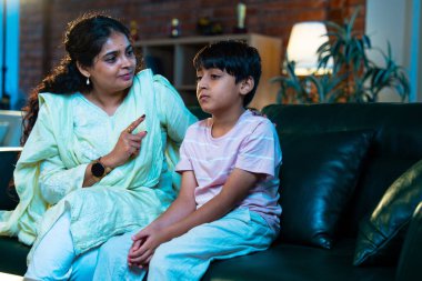 Hintli anne oğlunu azarlıyor, modern evde koltukta otururken, iyi alışkanlıklar öğretiyor, disiplini teşvik ediyor, kaliteli zaman geçirtiyor, çocuğa sevgiyle yol gösteriyor, ebeveyn çocuk ilişkisini kapalı alanda gösteriyor.