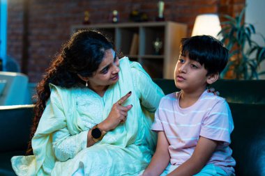 Hintli anne oğlunu azarlıyor, modern evde koltukta otururken, iyi alışkanlıklar öğretiyor, disiplini teşvik ediyor, kaliteli zaman geçirtiyor, çocuğa sevgiyle yol gösteriyor, ebeveyn çocuk ilişkisini kapalı alanda gösteriyor.