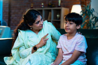 Hintli anne oğlunu azarlıyor, modern evde koltukta otururken, iyi alışkanlıklar öğretiyor, disiplini teşvik ediyor, kaliteli zaman geçirtiyor, çocuğa sevgiyle yol gösteriyor, ebeveyn çocuk ilişkisini kapalı alanda gösteriyor.