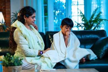 Hintli anne oğlunun ateşini kontrol ederken kanepenin üzerinde oturuyor, sağlığıyla ilgileniyor, sevgisini, sevgisini, sevgisini, aile bağlarını, evde aile yaşam tarzını, günlük rutini besliyor.