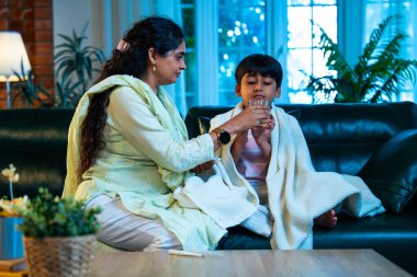 Hintli anne oğlunun ateşini kontrol ederken kanepenin üzerinde oturuyor, sağlığıyla ilgileniyor, sevgisini, sevgisini, sevgisini, aile bağlarını, evde aile yaşam tarzını, günlük rutini besliyor.