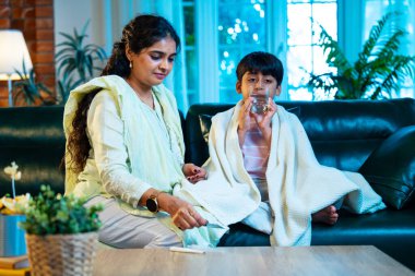 Hintli anne oğlunun ateşini kontrol ederken kanepenin üzerinde oturuyor, sağlığıyla ilgileniyor, sevgisini, sevgisini, sevgisini, aile bağlarını, evde aile yaşam tarzını, günlük rutini besliyor.