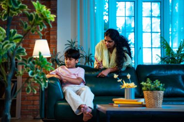 Hintli anne oğlunu azarlıyor, modern evde koltukta otururken, iyi alışkanlıklar öğretiyor, disiplini teşvik ediyor, kaliteli zaman geçirtiyor, çocuğa sevgiyle yol gösteriyor, ebeveyn çocuk ilişkisini kapalı alanda gösteriyor.