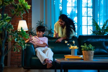 Hintli anne oğlunu azarlıyor, modern evde koltukta otururken, iyi alışkanlıklar öğretiyor, disiplini teşvik ediyor, kaliteli zaman geçirtiyor, çocuğa sevgiyle yol gösteriyor, ebeveyn çocuk ilişkisini kapalı alanda gösteriyor.