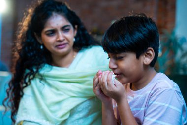 Hintli küçük bir çocuk modern bir evde annesinin yanında otururken öksürüyor, viral semptomlar gösteriyor, kendini iyi hissetmiyor, annesinin bakımı, çocuk bakımı, aile bağı, sağlık farkındalığı, bakımı,