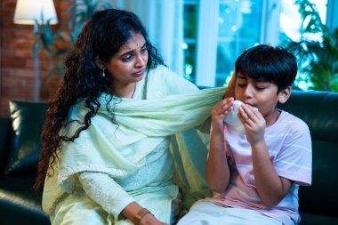 Hintli küçük bir çocuk modern bir evde annesinin yanında otururken öksürüyor, viral semptomlar gösteriyor, kendini iyi hissetmiyor, annesinin bakımı, çocuk bakımı, aile bağı, sağlık farkındalığı, bakımı,