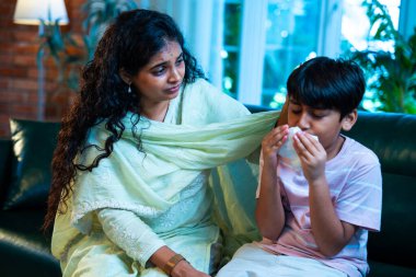 Hintli küçük bir çocuk modern bir evde annesinin yanında otururken öksürüyor, viral semptomlar gösteriyor, kendini iyi hissetmiyor, annesinin bakımı, çocuk bakımı, aile bağı, sağlık farkındalığı, bakımı,