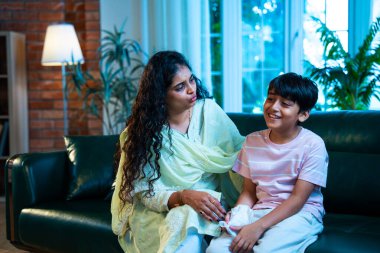 Hintli küçük bir çocuk modern bir evde annesinin yanında otururken öksürüyor, viral semptomlar gösteriyor, kendini iyi hissetmiyor, annesinin bakımı, çocuk bakımı, aile bağı, sağlık farkındalığı, bakımı,