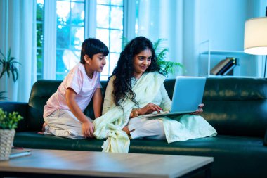 Hintli küçük çocuk ve anne internette sörf yapıyor ya da internetten alışveriş yapıyor modern evdeki kanepede otururken, birlikte kaliteli zaman geçiriyorlar, bağlanma, bağ kurma, ebeveyn çocuk yaşam tarzı gösteriyorlar.