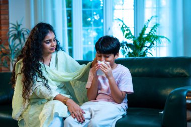 Hintli küçük bir çocuk modern bir evde annesinin yanında otururken öksürüyor, viral semptomlar gösteriyor, kendini iyi hissetmiyor, annesinin bakımı, çocuk bakımı, aile bağı, sağlık farkındalığı, bakımı,