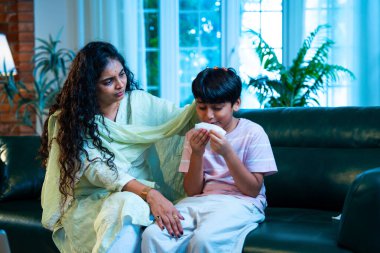 Hintli küçük bir çocuk modern bir evde annesinin yanında otururken öksürüyor, viral semptomlar gösteriyor, kendini iyi hissetmiyor, annesinin bakımı, çocuk bakımı, aile bağı, sağlık farkındalığı, bakımı,