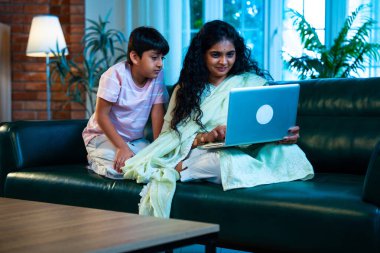 Hintli küçük çocuk ve anne internette sörf yapıyor ya da internetten alışveriş yapıyor modern evdeki kanepede otururken, birlikte kaliteli zaman geçiriyorlar, bağlanma, bağ kurma, ebeveyn çocuk yaşam tarzı gösteriyorlar.