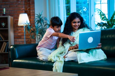 Hintli küçük çocuk ve anne internette sörf yapıyor ya da internetten alışveriş yapıyor modern evdeki kanepede otururken, birlikte kaliteli zaman geçiriyorlar, bağlanma, bağ kurma, ebeveyn çocuk yaşam tarzı gösteriyorlar.