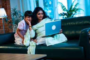 Hintli küçük çocuk ve anne internette sörf yapıyor ya da internetten alışveriş yapıyor modern evdeki kanepede otururken, birlikte kaliteli zaman geçiriyorlar, bağlanma, bağ kurma, ebeveyn çocuk yaşam tarzı gösteriyorlar.