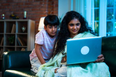 Hintli küçük çocuk ve anne internette sörf yapıyor ya da internetten alışveriş yapıyor modern evdeki kanepede otururken, birlikte kaliteli zaman geçiriyorlar, bağlanma, bağ kurma, ebeveyn çocuk yaşam tarzı gösteriyorlar.