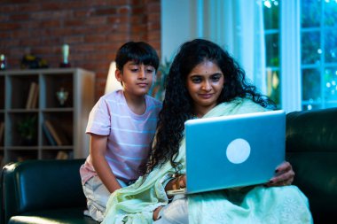 Hintli küçük çocuk ve anne internette sörf yapıyor ya da internetten alışveriş yapıyor modern evdeki kanepede otururken, birlikte kaliteli zaman geçiriyorlar, bağlanma, bağ kurma, ebeveyn çocuk yaşam tarzı gösteriyorlar.