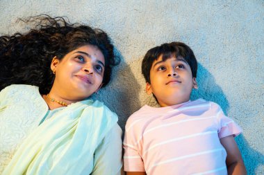 Hintli anne oğul modern evde halıda yatarken konuşuyor, birlikte kaliteli zaman geçiriyorlar, sevgi, neşe, sıcaklık, bağ, ebeveyn çocuk ilişkisi, kapalı alanda aile yaşam tarzı paylaşıyorlar.