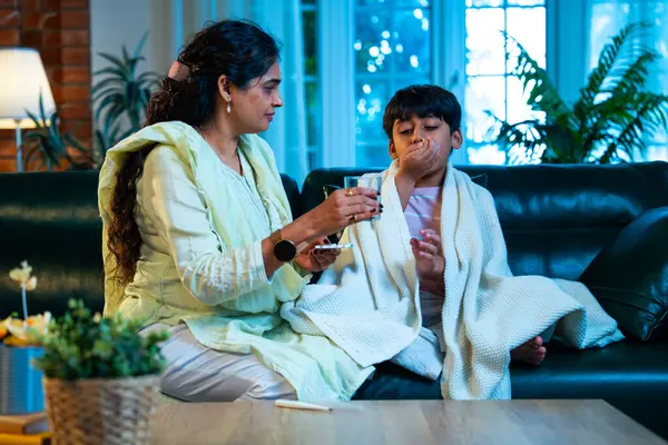 Hintli anne oğlunun ateşini kontrol ederken kanepenin üzerinde oturuyor, sağlığıyla ilgileniyor, sevgisini, sevgisini, sevgisini, aile bağlarını, evde aile yaşam tarzını, günlük rutini besliyor.