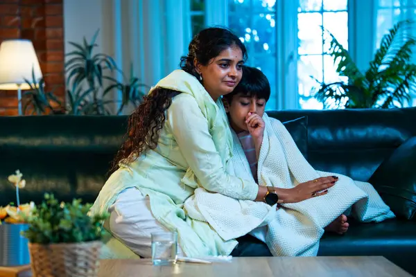 Hintli anne oğlunun ateşini kontrol ederken kanepenin üzerinde oturuyor, sağlığıyla ilgileniyor, sevgisini, sevgisini, sevgisini, aile bağlarını, evde aile yaşam tarzını, günlük rutini besliyor.