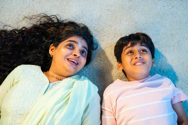 Hintli anne oğul modern evde halıda yatarken konuşuyor, birlikte kaliteli zaman geçiriyorlar, sevgi, neşe, sıcaklık, bağ, ebeveyn çocuk ilişkisi, kapalı alanda aile yaşam tarzı paylaşıyorlar.